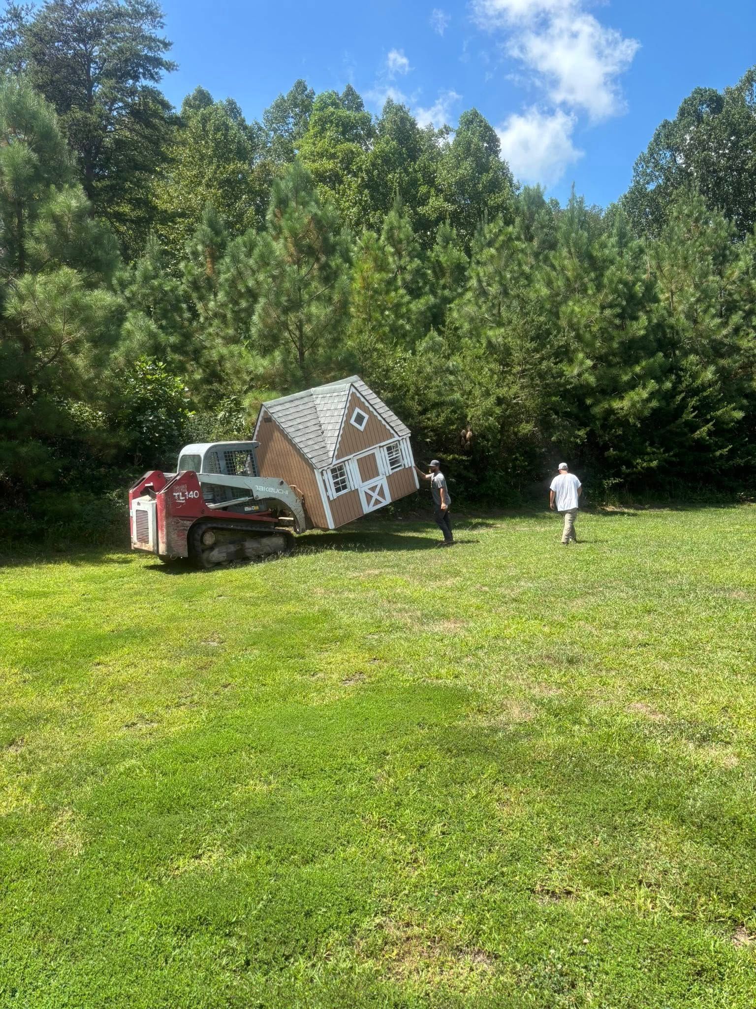 A red skid steer loader lifts a small, decorative wooden house in a grassy field near a line of trees.