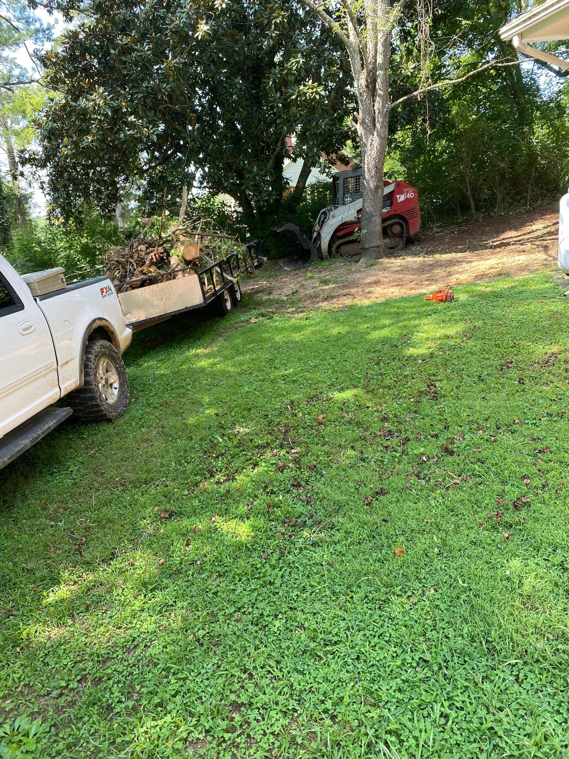 A white pickup truck with a trailer loaded with debris parked on a lawn near a red skid steer working by a tree line.