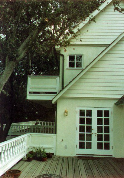 A two-story white house with a deck, a balcony, French doors, and a tree nearby under an overcast sky.