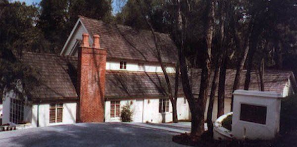 A white house with a brick chimney and a dark shingled roof, surrounded by trees on a paved lot.