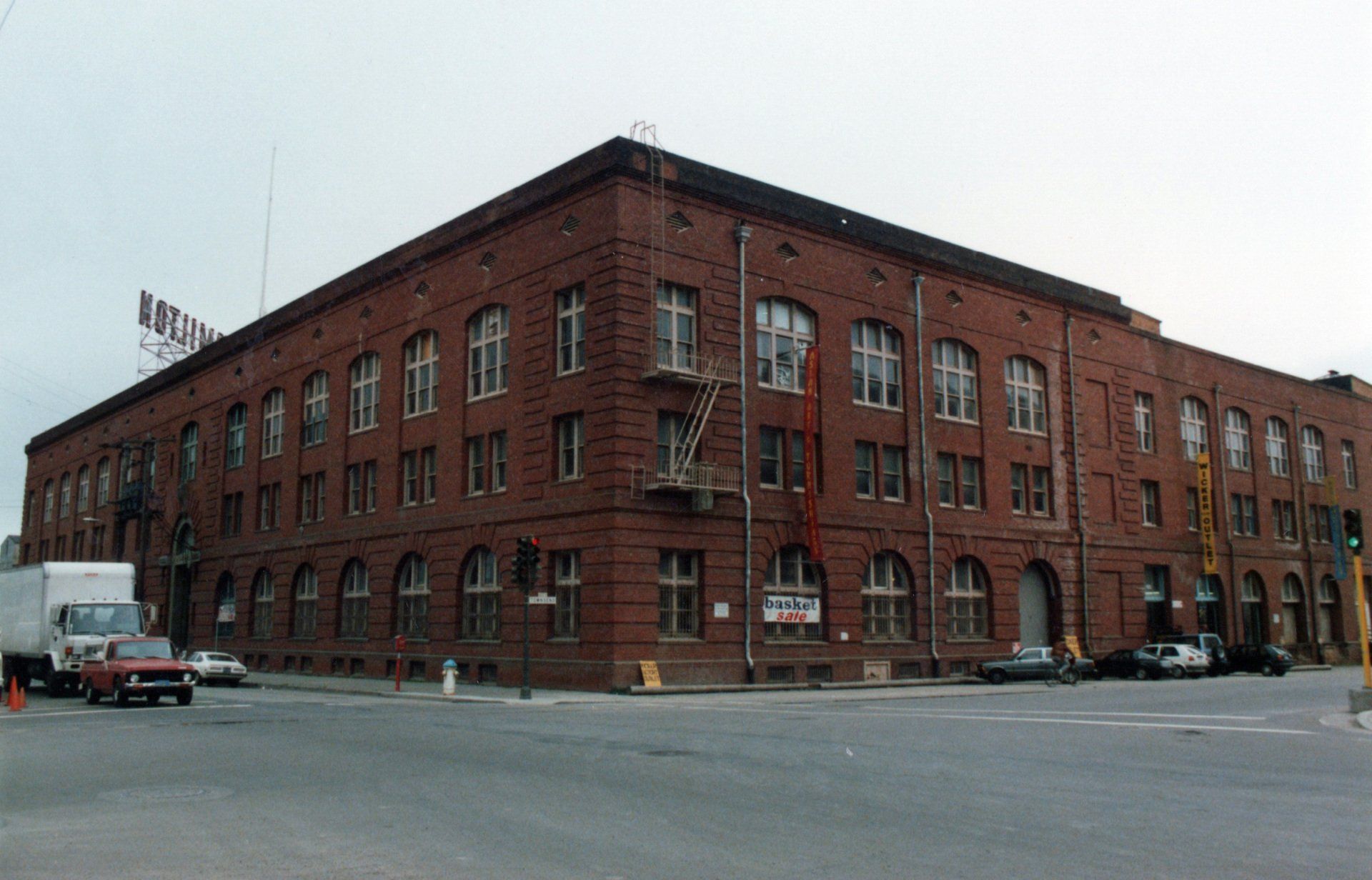 A large, red brick building with an arched ground floor and a rooftop sign at a street corner on an overcast day.