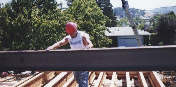 A construction worker wearing a red hard hat guides a large steel beam over an open wooden floor frame.