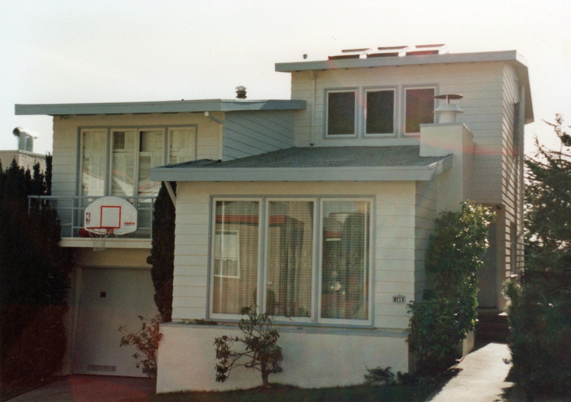 A two-story white house with a flat roof, large front windows, an attached garage, and a basketball hoop on the balcony.