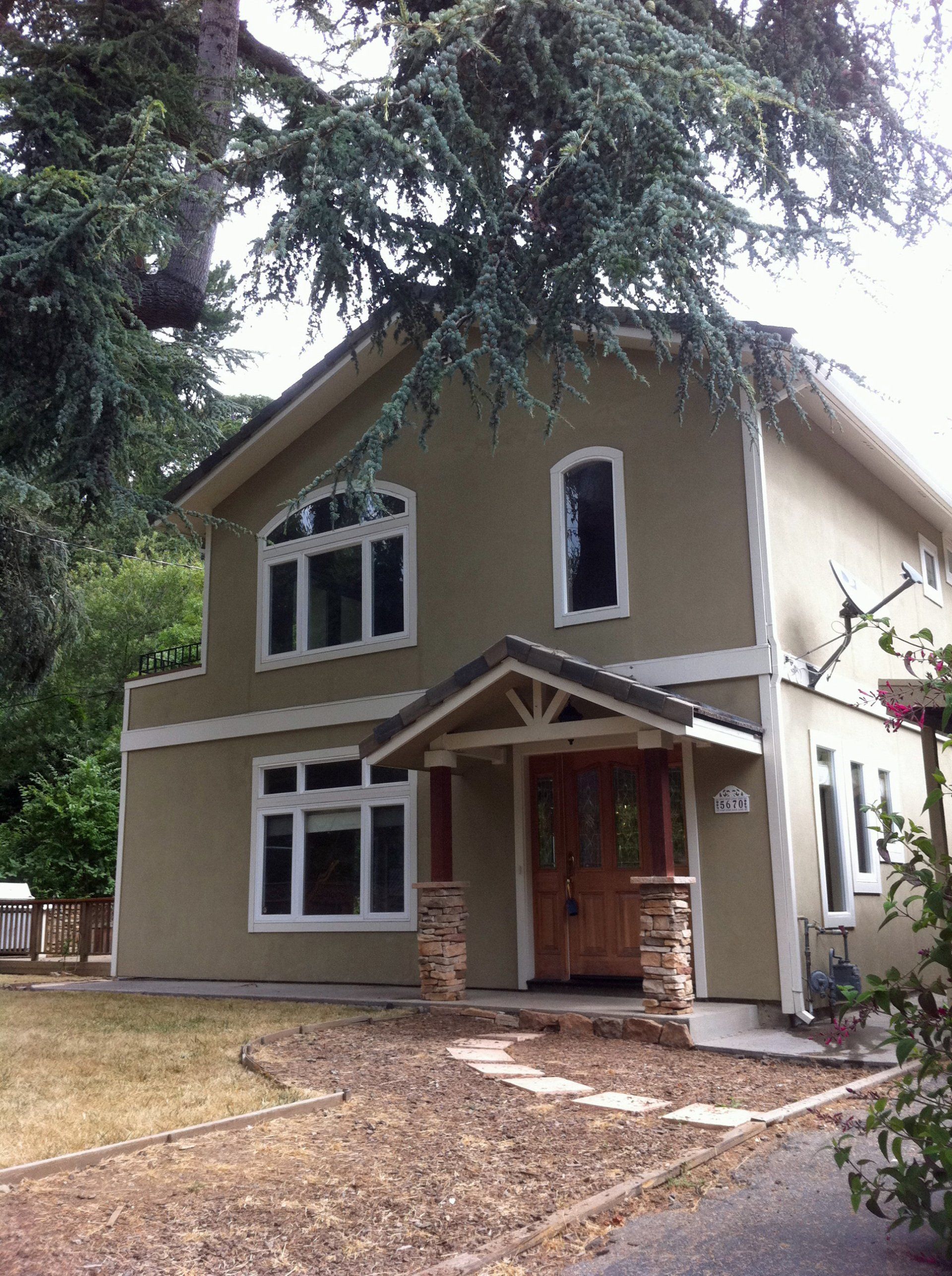 A two-story, tan-colored house with a peaked roof, stone porch pillars, a wooden front door, and a large shade tree.