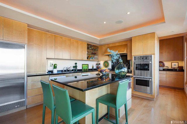 Modern kitchen with light wood cabinets, black granite countertops, an island with two green chairs, and stainless ovens.