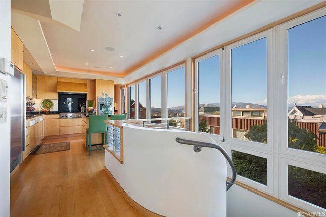 A modern kitchen with light wood cabinets, green chairs, and a white curved wall, overlooking large windows with a view.