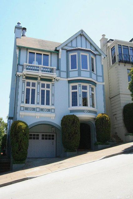 A light blue, multi-story house with a garage and bay windows, located on a steep, paved street in a residential area.