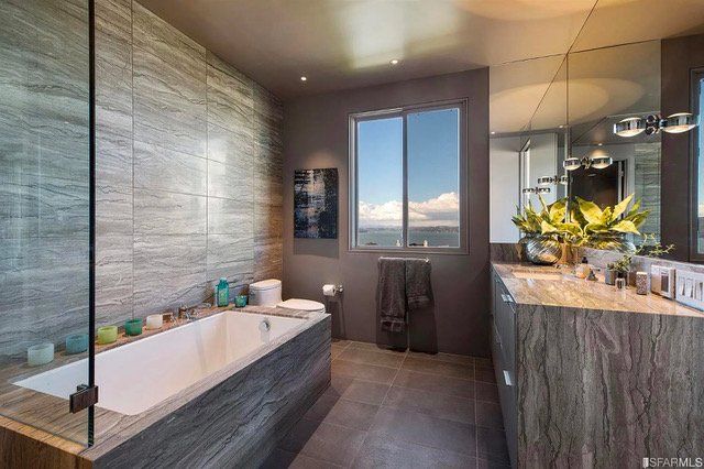 A modern bathroom with a white soaking tub, wood-grain tiled walls, a vanity with a large mirror, and a window with a view.