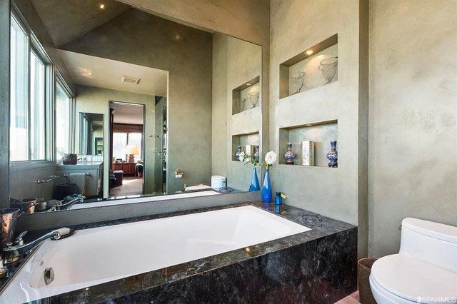 A bathroom featuring a white bathtub encased in dark marble, a large mirror, and wall niches holding decorative items.