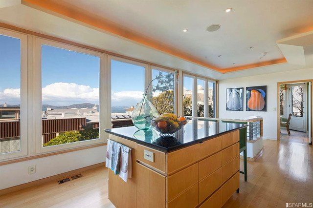 A modern kitchen island with a dark countertop and wood cabinets sits before a bright room with large glass windows.