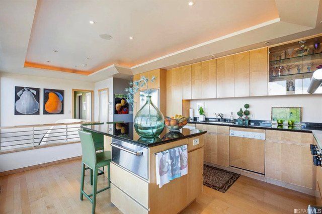 A modern kitchen with light wood cabinets, a black granite island with green chairs, and orange accents in wall art.
