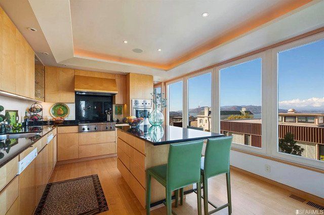 Modern kitchen with light wood cabinetry, a granite-topped island with two green stools, and floor-to-ceiling windows.