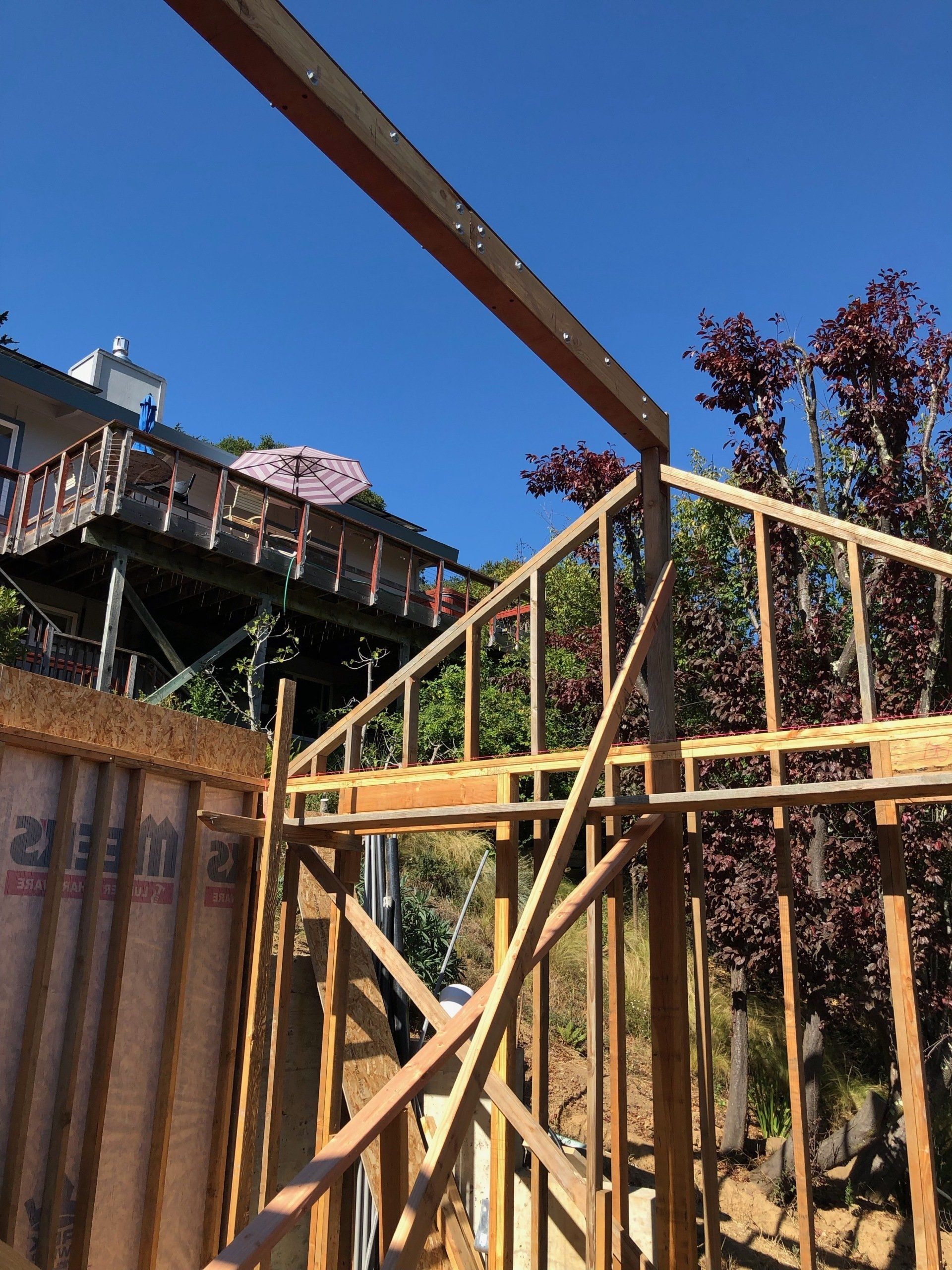 A wooden framed wall and roof structure under construction against a clear blue sky, with a house visible on a hillside.