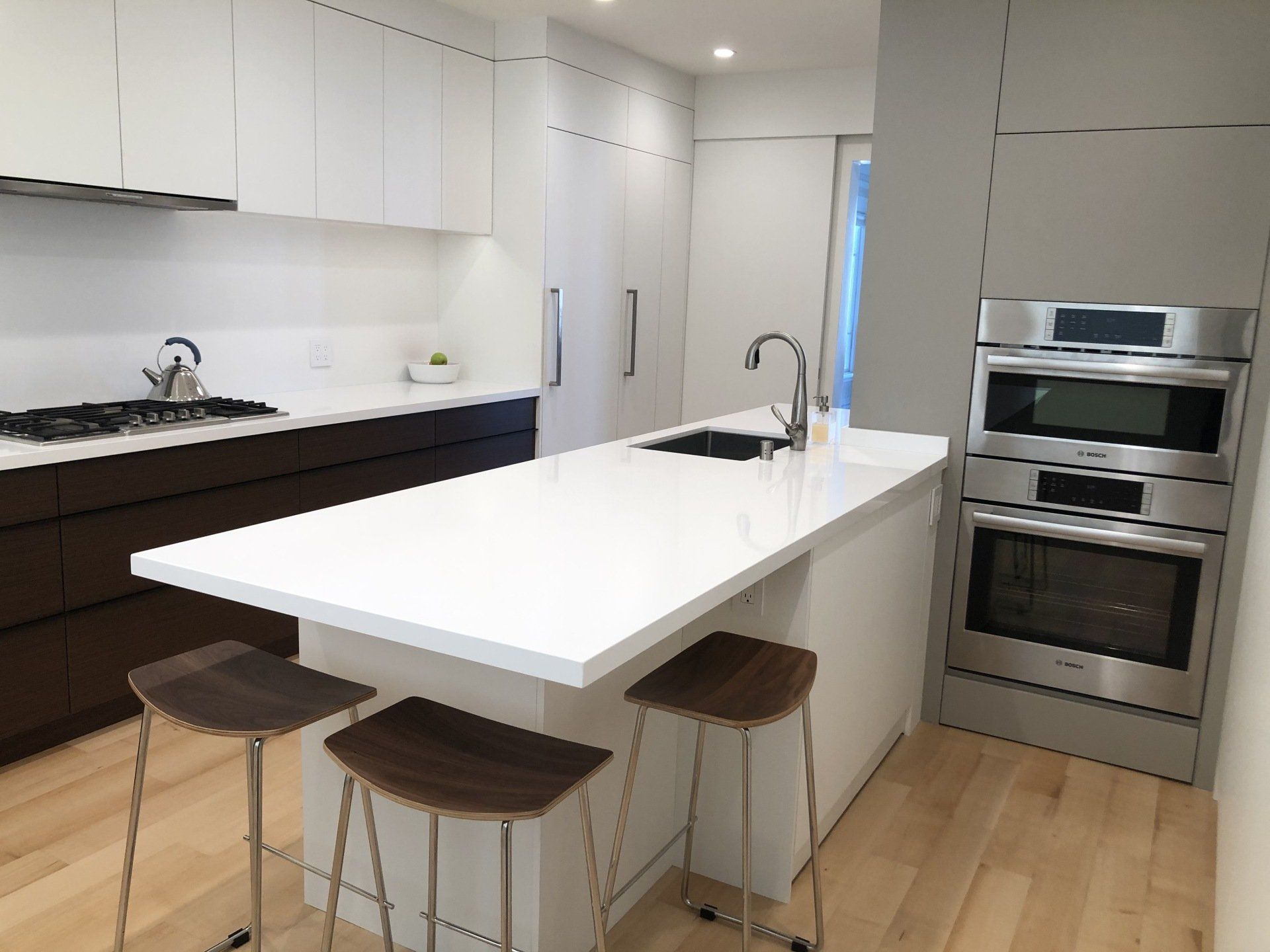 A modern kitchen with a white island, wooden stools, dark cabinetry, white upper cabinets, and a stainless steel oven.