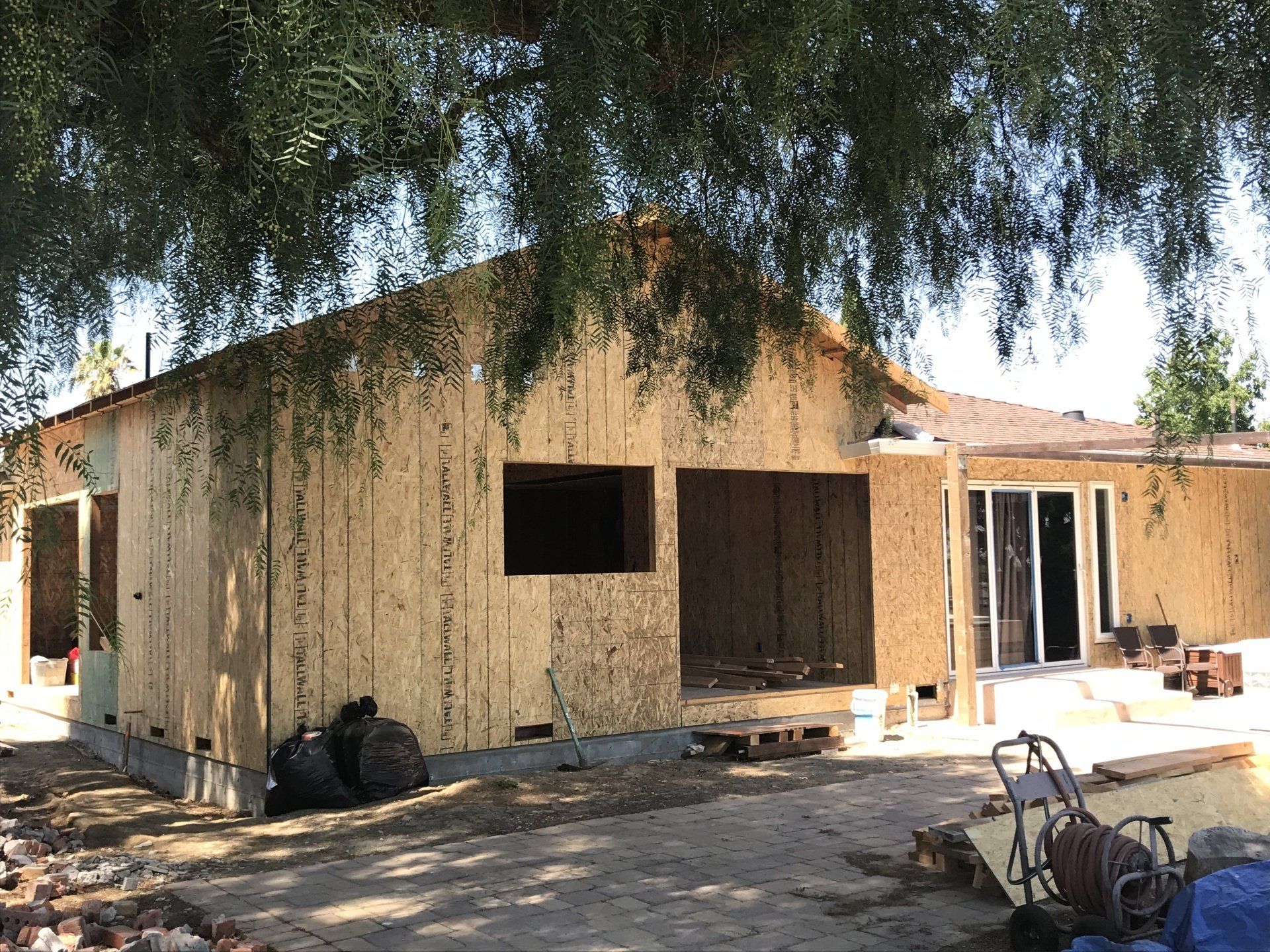 A house exterior under construction, featuring bare plywood walls, window cutouts, and a patio area shaded by a tree.