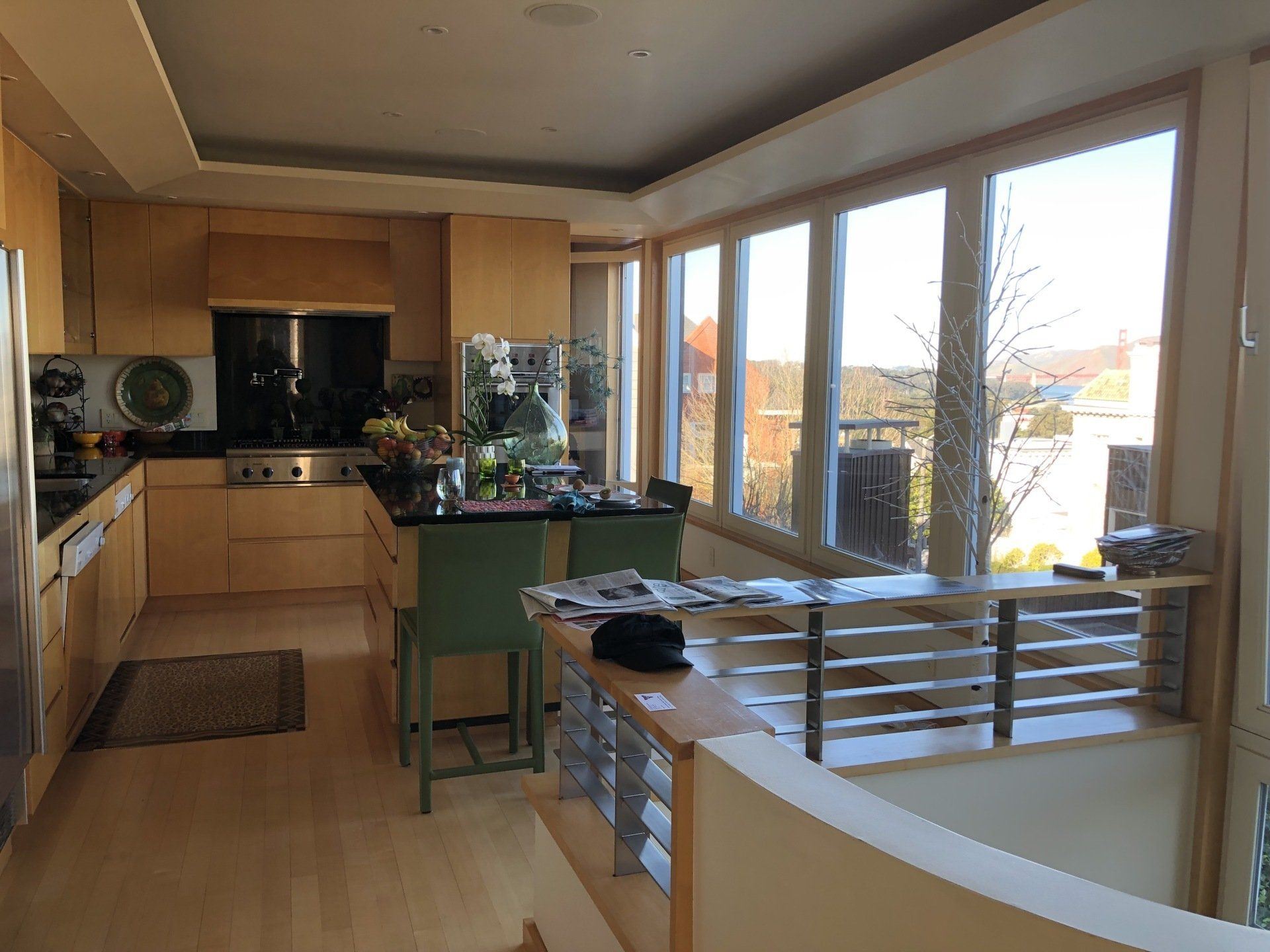 A modern kitchen with light wood cabinets, black countertops, an island with green chairs, and large windows with a view.