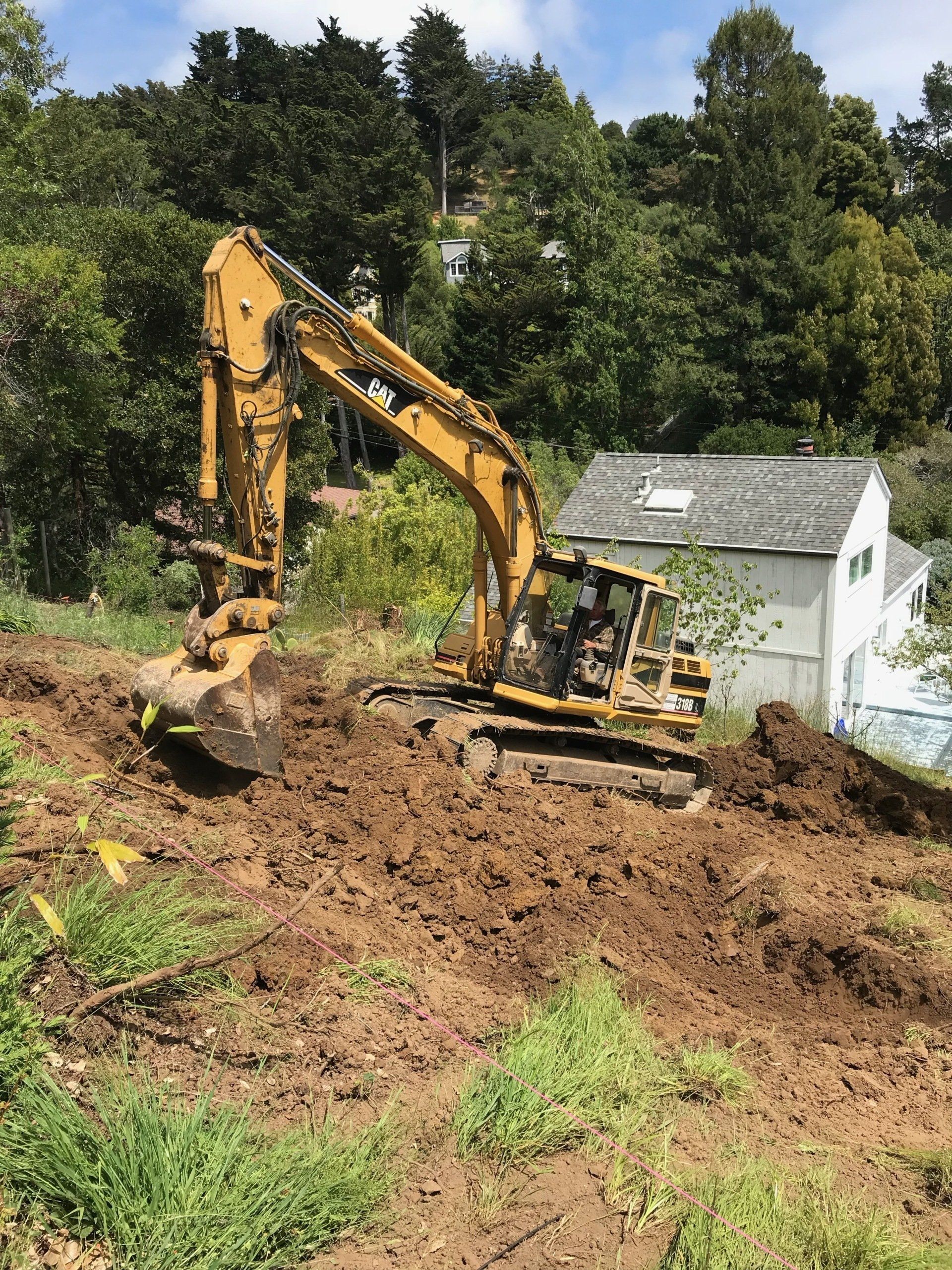 A yellow excavator sits on a dirt hillside, digging into the earth above a white house and trees.