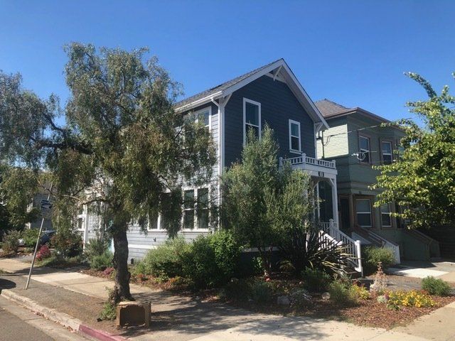 A two-story dark blue suburban house with white trim and a small front porch, flanked by trees and greenery under blue sky.