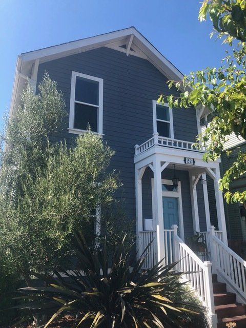 A two-story blue house with white trim, a front porch, and a staircase, framed by a tree and a large spiky plant.