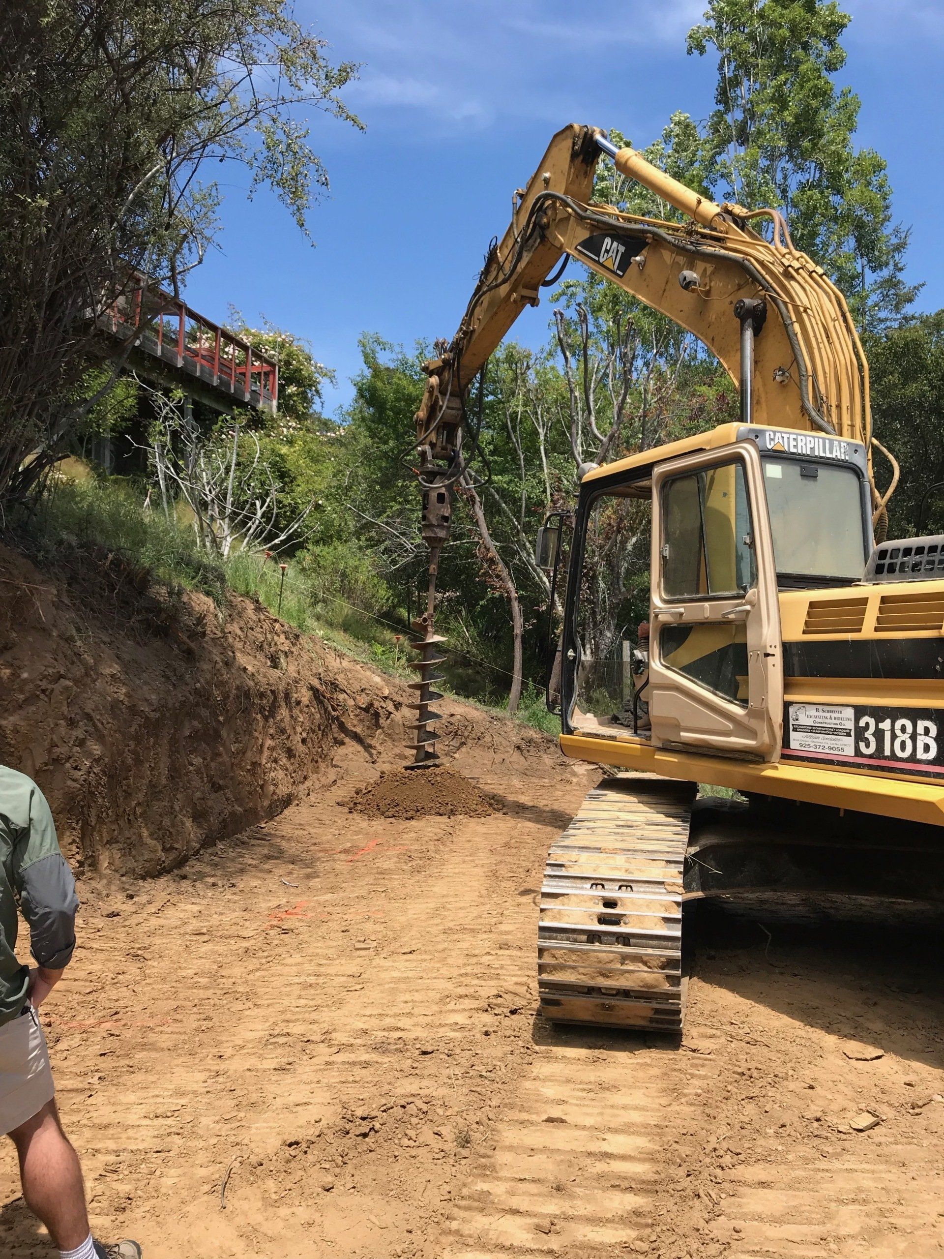 A yellow Caterpillar 318B excavator drills a hole into a dirt slope near a wooded area.