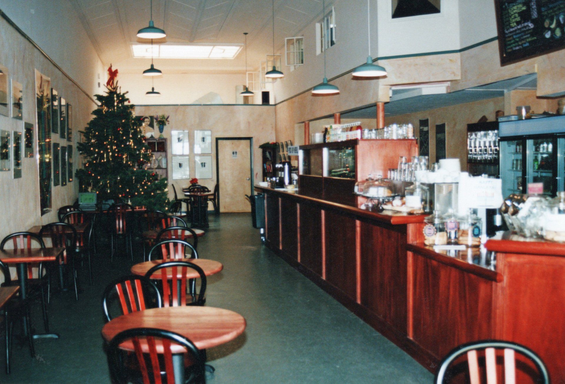 A cafe interior featuring wooden tables, chairs, a long serving counter, and a Christmas tree in the corner.