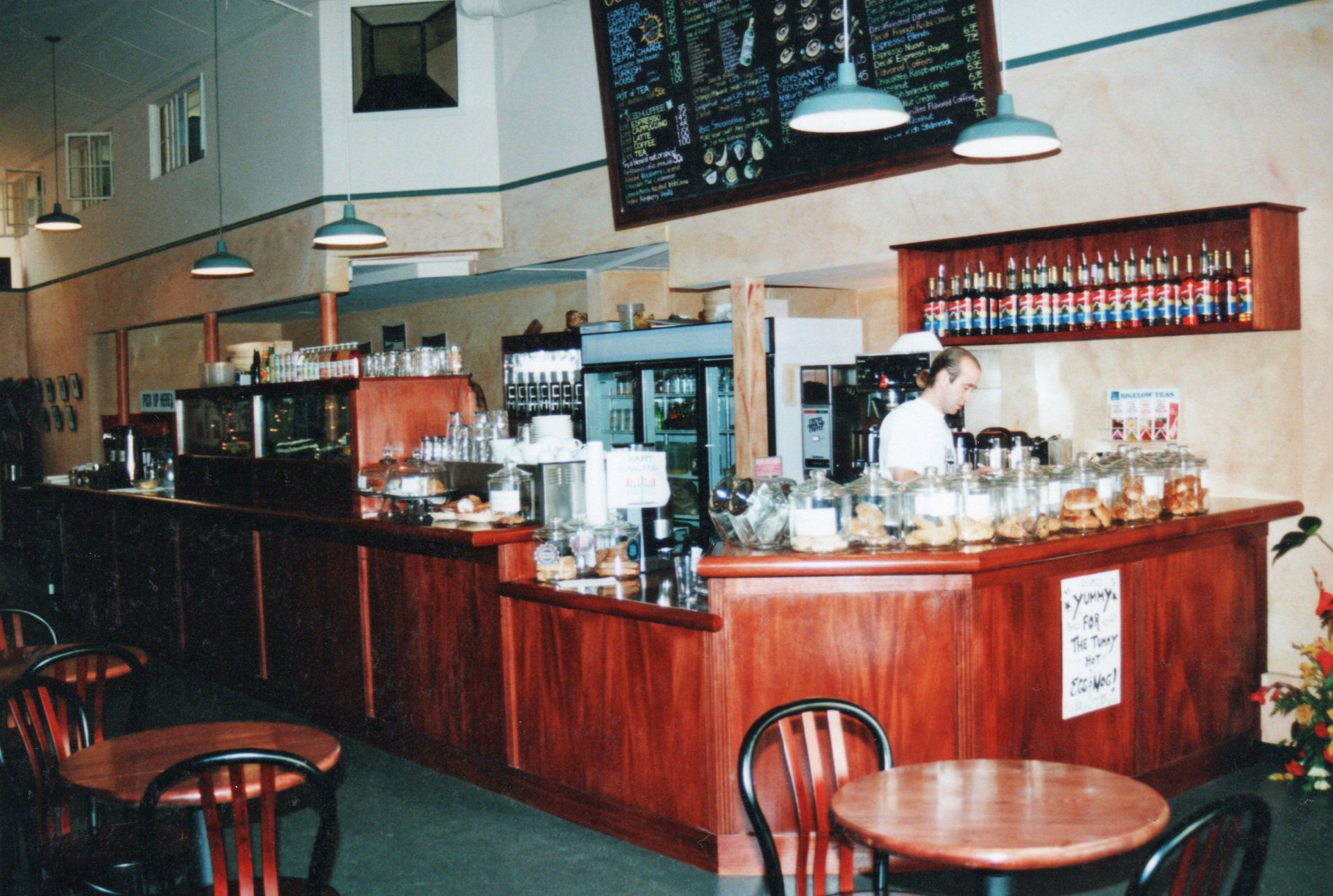 A coffee shop interior features a wooden counter, a person working behind it, a menu board, and tables with chairs.
