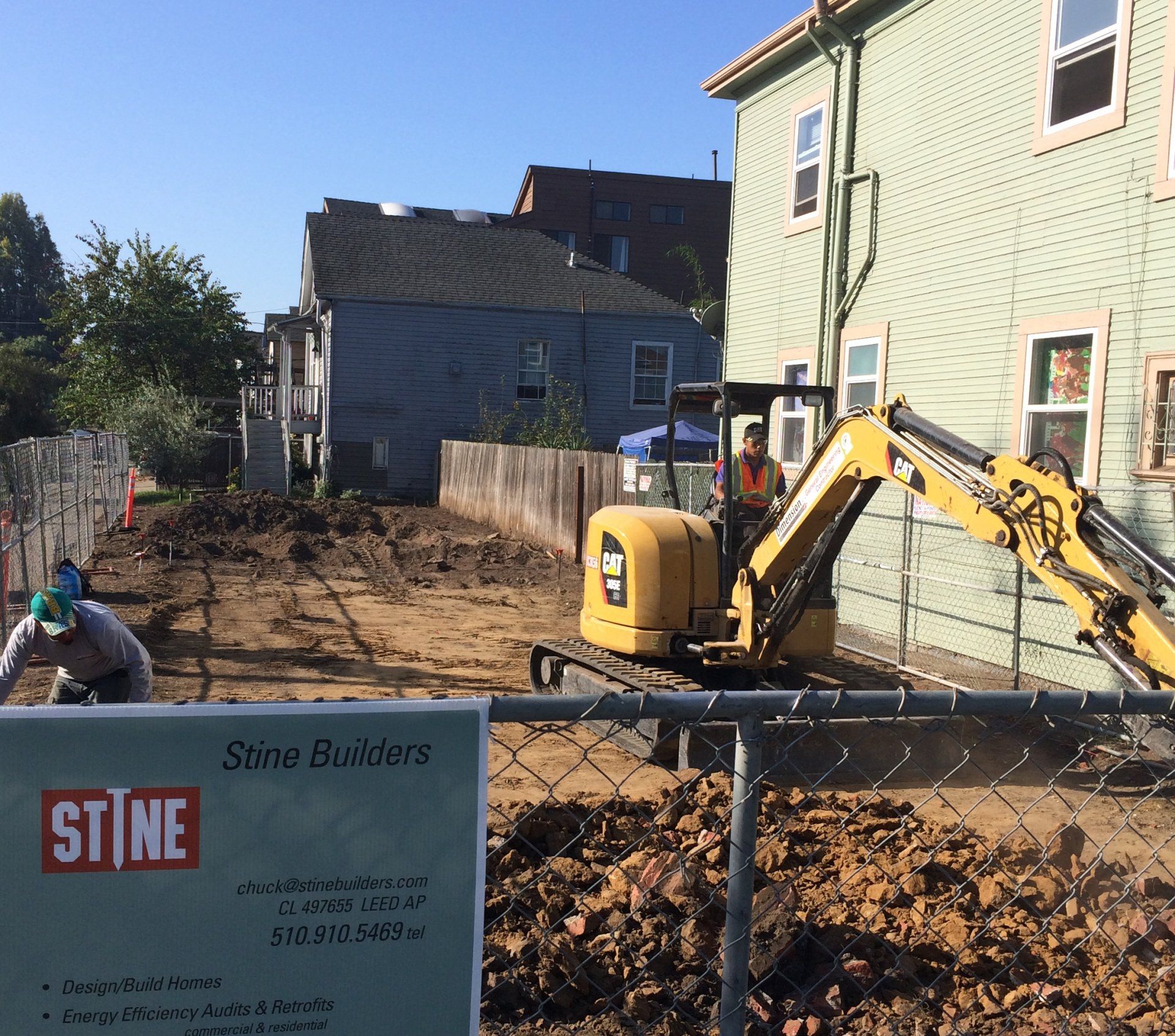 A yellow Caterpillar excavator operates on a dirt construction site next to a building, with a Stine Builders sign nearby.