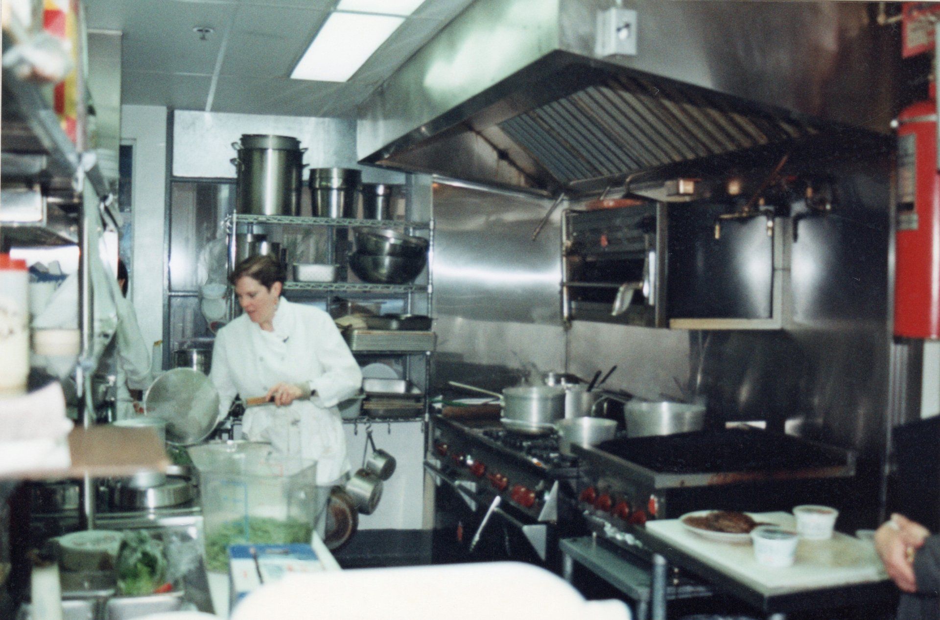 A professional kitchen with a chef cooking at a stove under a metal exhaust hood, surrounded by pots and prep stations.