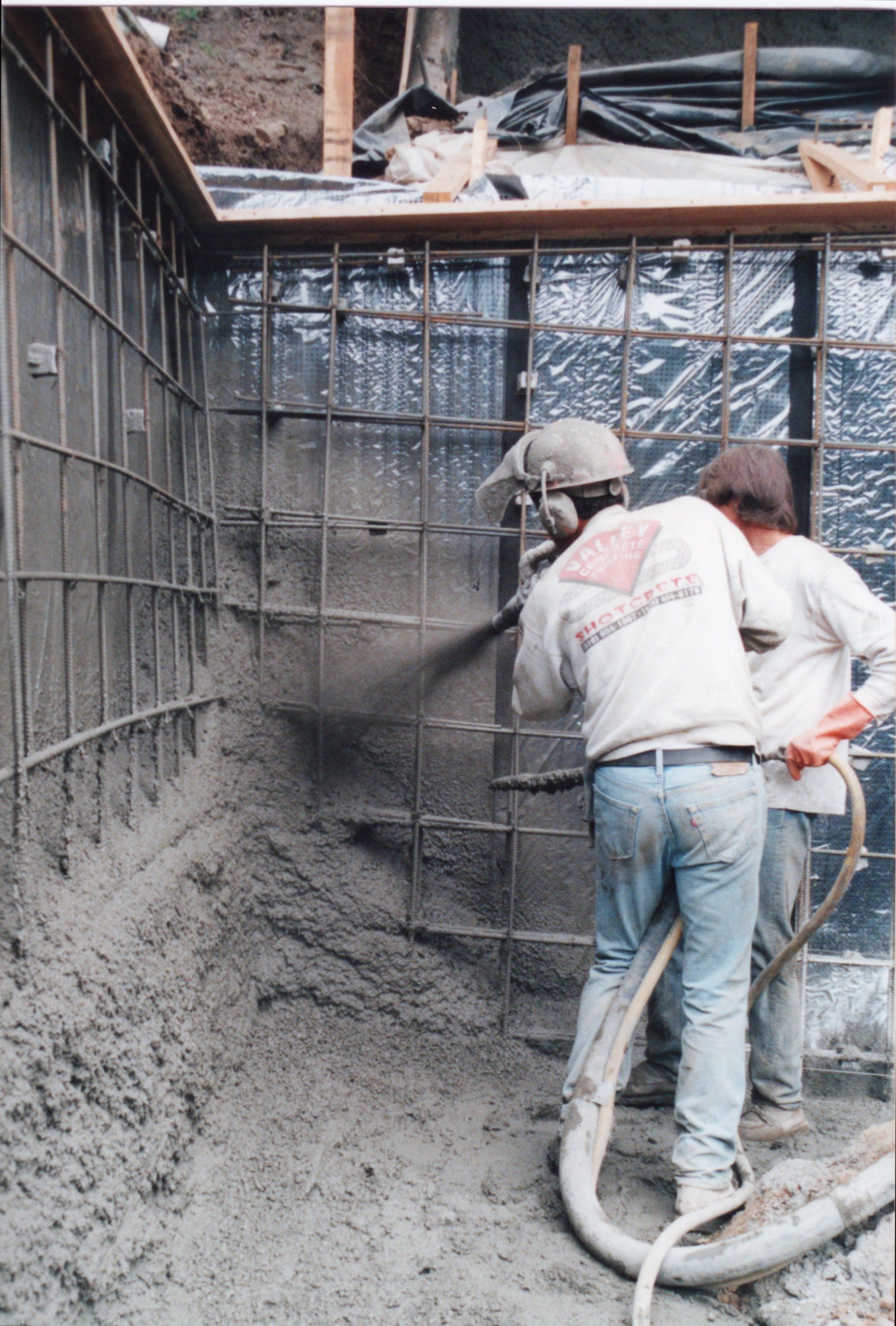 Two construction workers use a pressurized hose to spray concrete onto a steel-reinforced wall in an open excavation site.