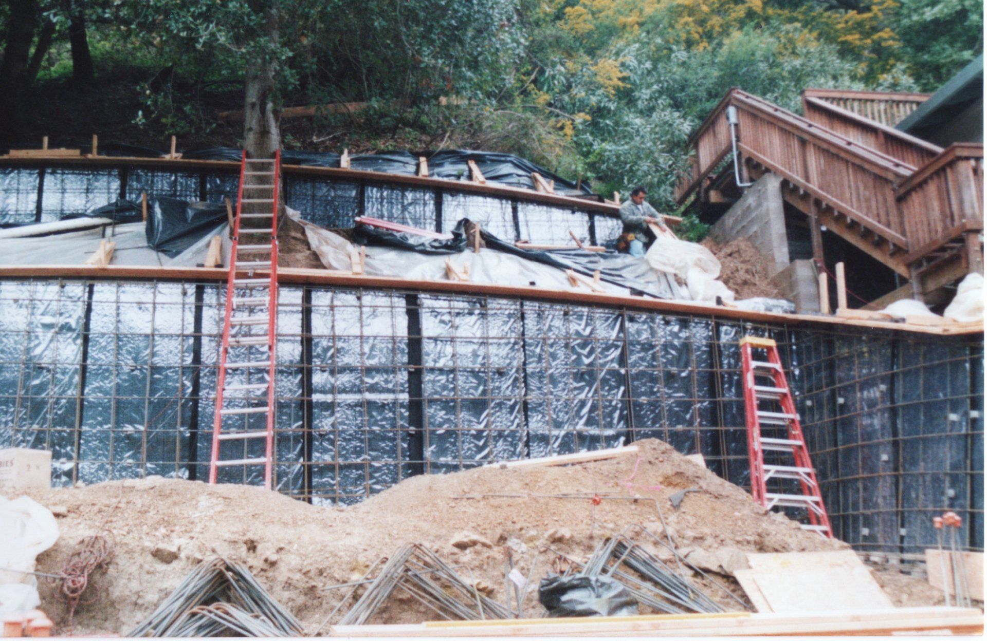 Construction workers install steel reinforcement and waterproofing material for a concrete retaining wall on a hillside.