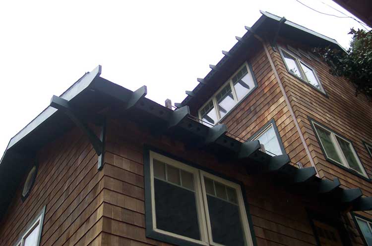 A low-angle view of a wooden house exterior with shingled walls, dark trim, and decorative roof rafter tails.