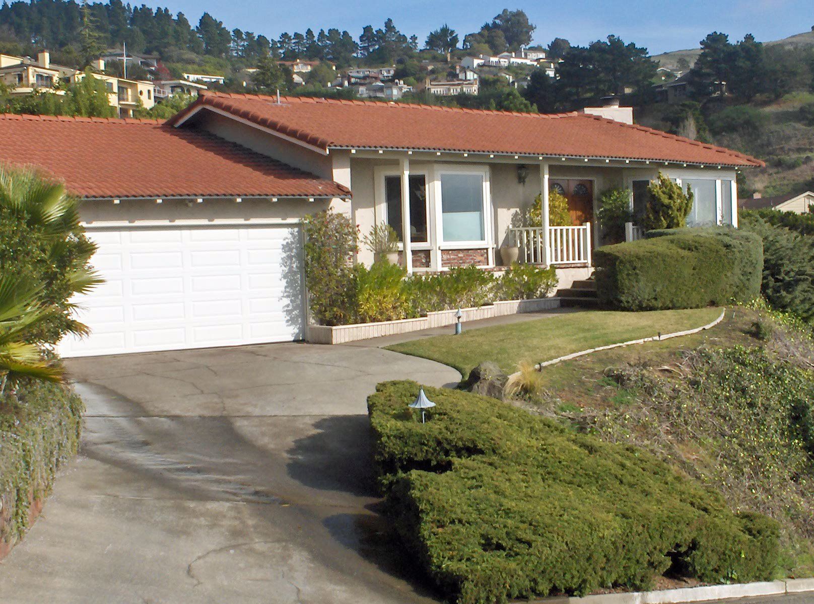 A one-story suburban home with a red tile roof, white garage door, and manicured green landscaping on a sunny day.