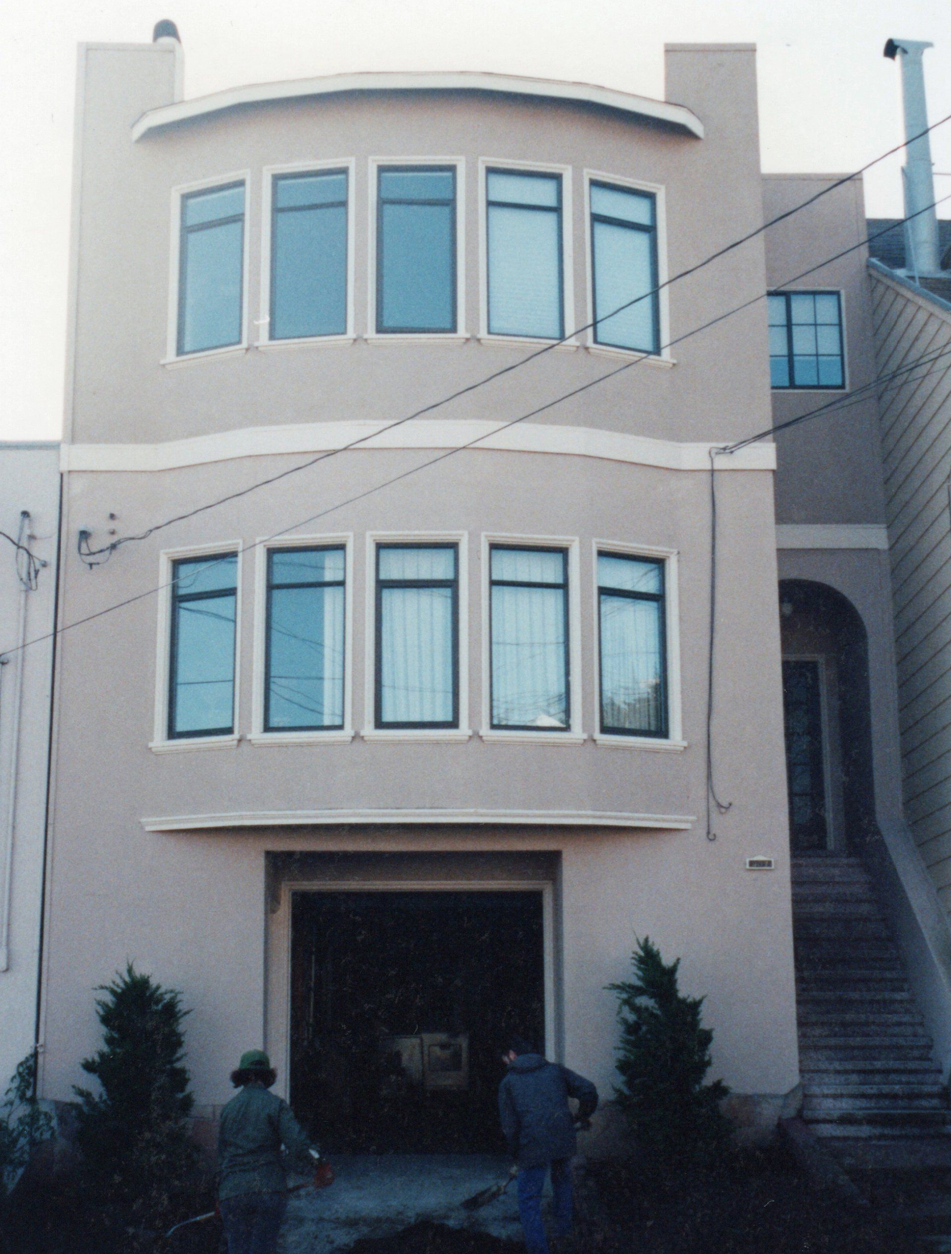A light-colored, two-story house with bay windows and a garage, featuring two individuals standing at the entrance.
