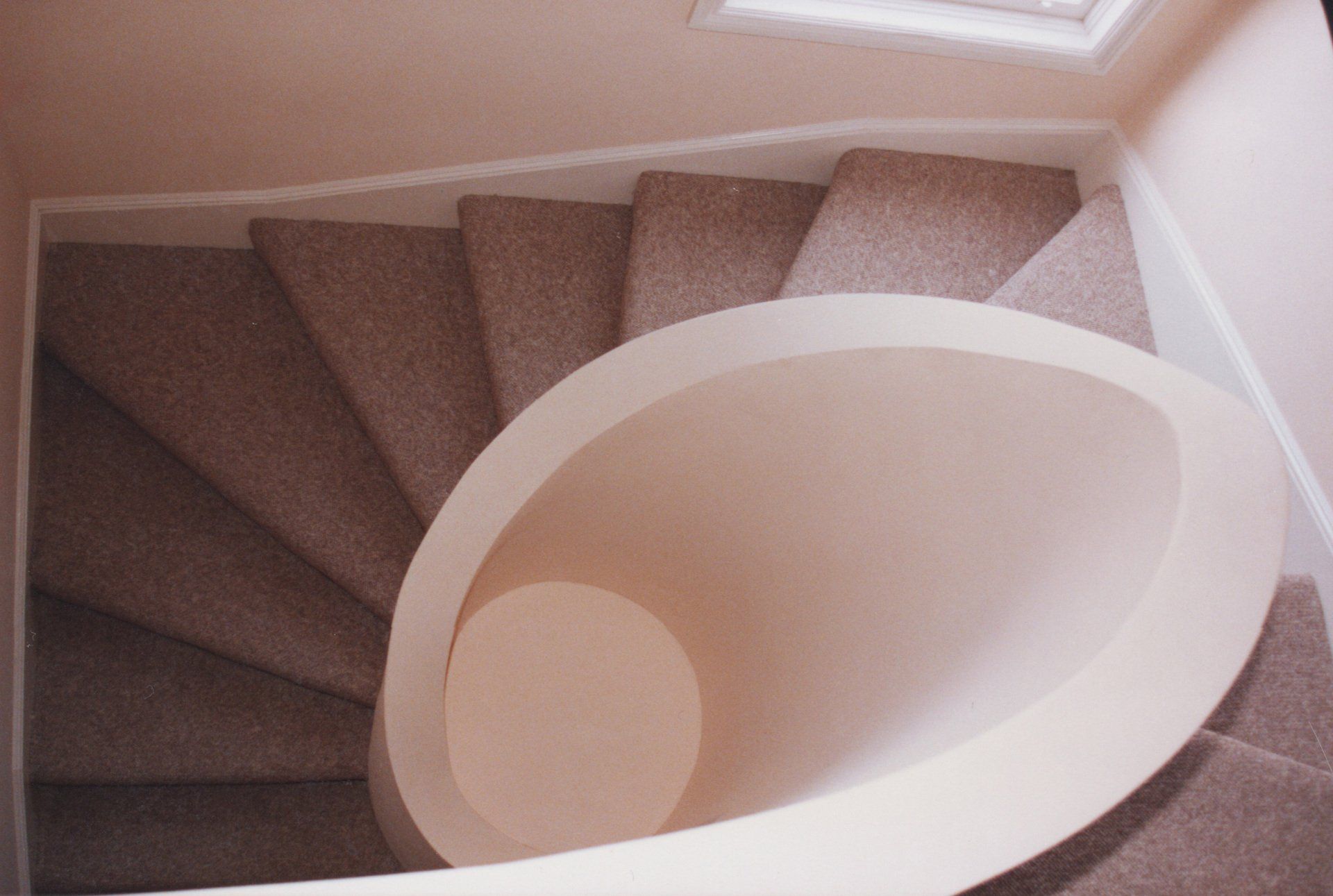 An overhead view of a curved staircase with tan carpet winding around a smooth, elliptical white wall opening.