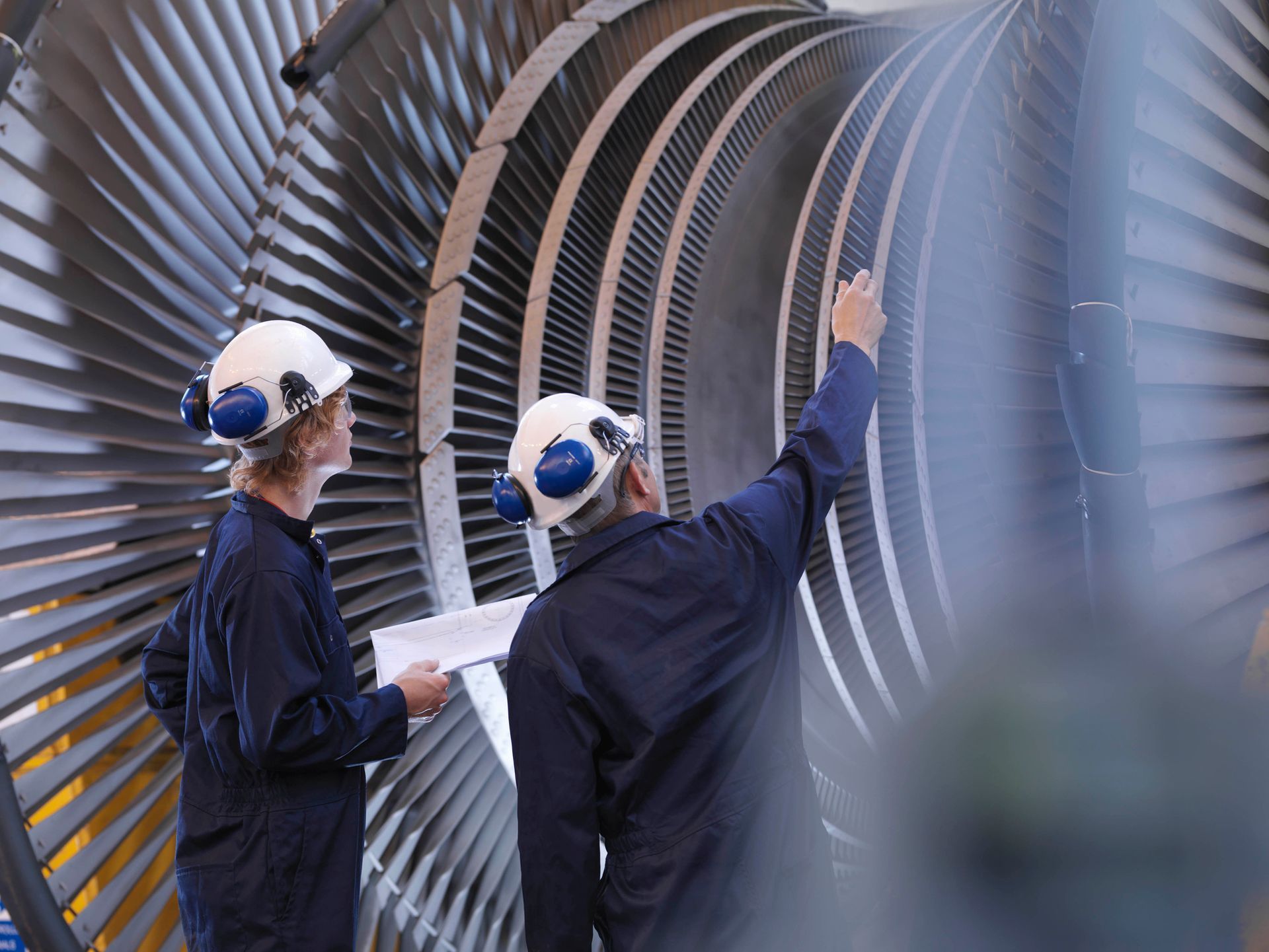 Due ingegneri in tuta blu e casco ispezionano una grande turbina con pale argentate.