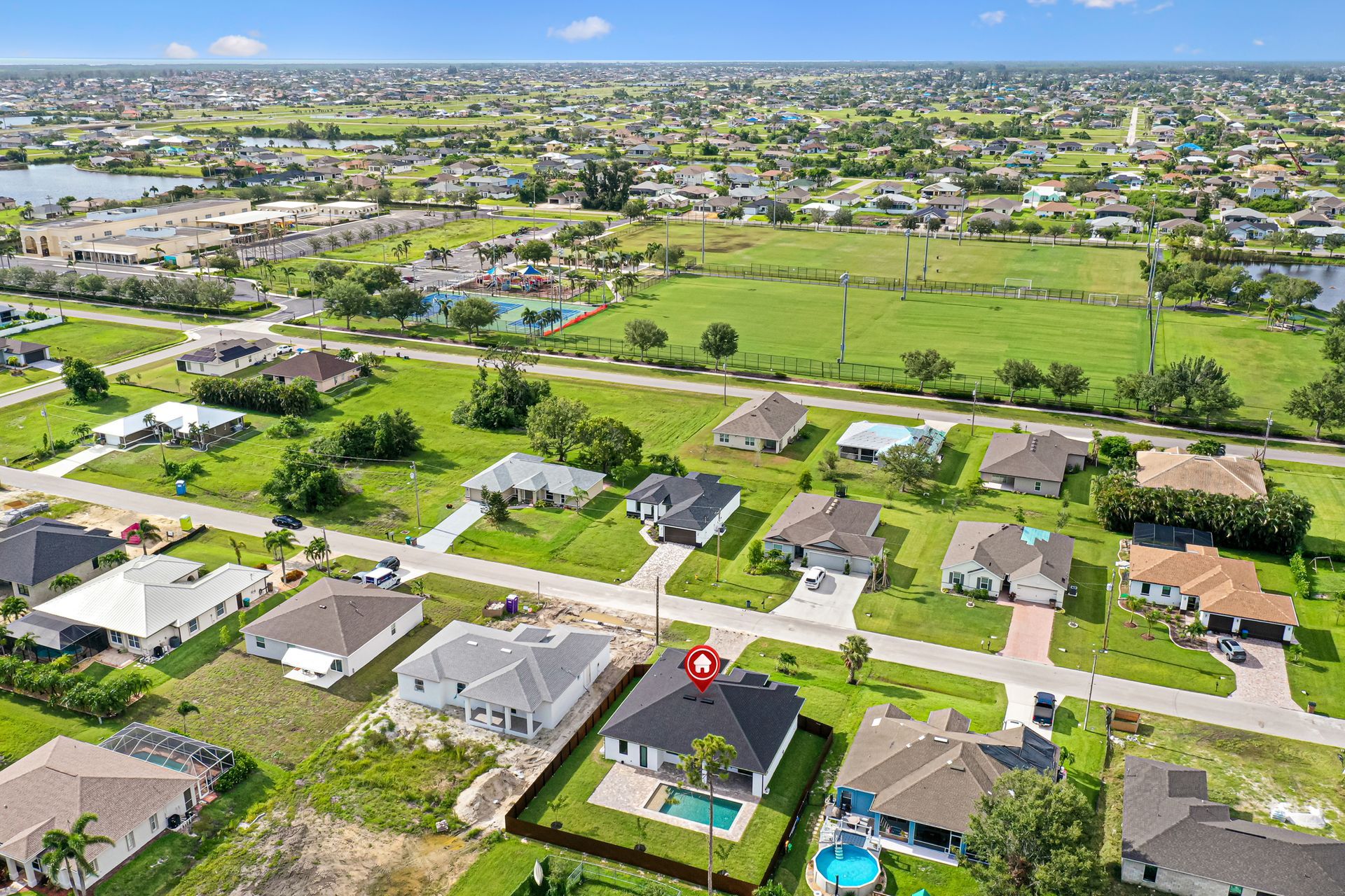 Aerial view of suburban homes, green lawns, and a small pool in a sunny neighborhood.