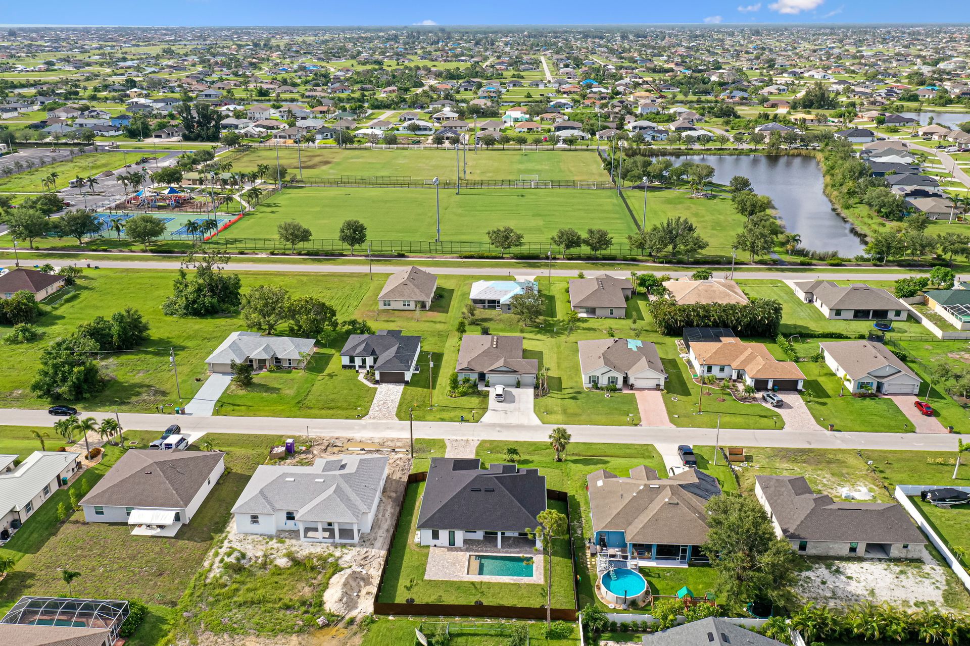 Aerial view of suburban homes with pools and green spaces, near a large field and canal.