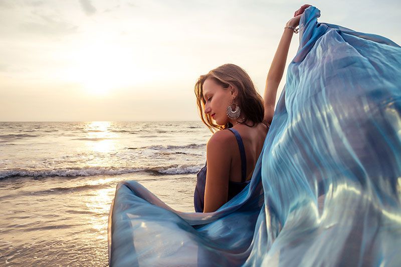 Virtual Dispensary - Beautiful brunette girl in blue gray chameleon dress with long train sitting on a beach at sunset