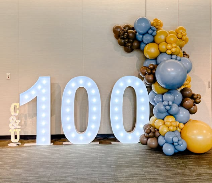 The number 100 is surrounded by balloons and lights
