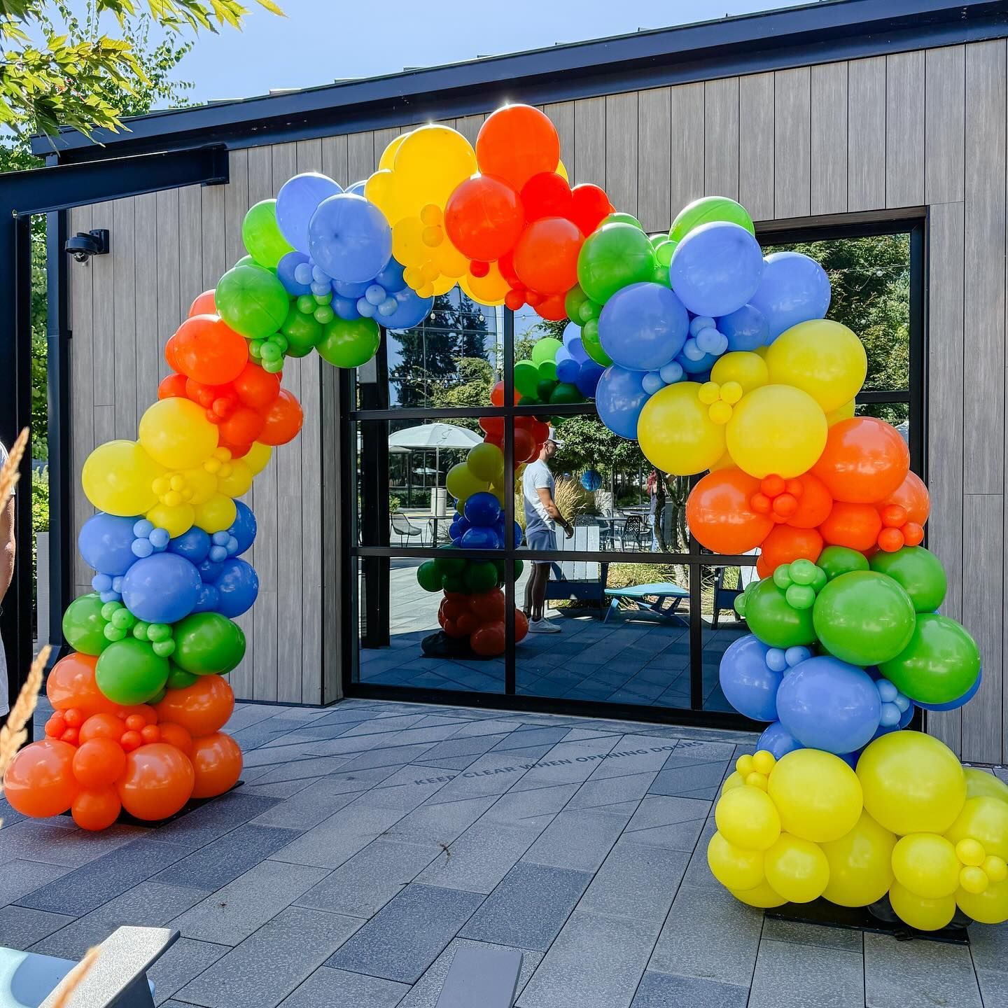 Rainbow balloon archway at a glass door entrance, on a stone patio.
