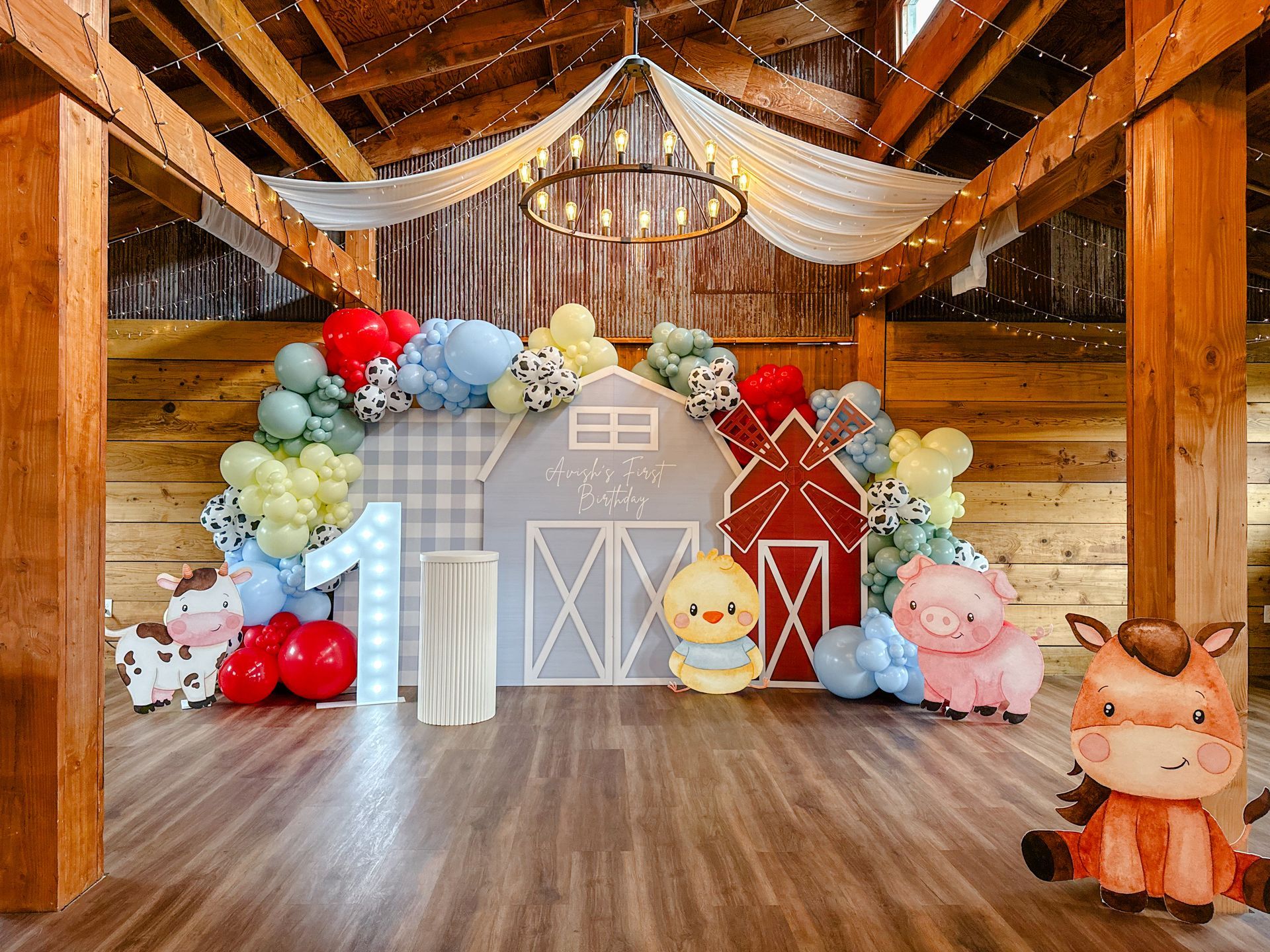 Farm-themed first birthday party setup in a barn, featuring balloon arches, a barn backdrop, and cute animal illustrations.