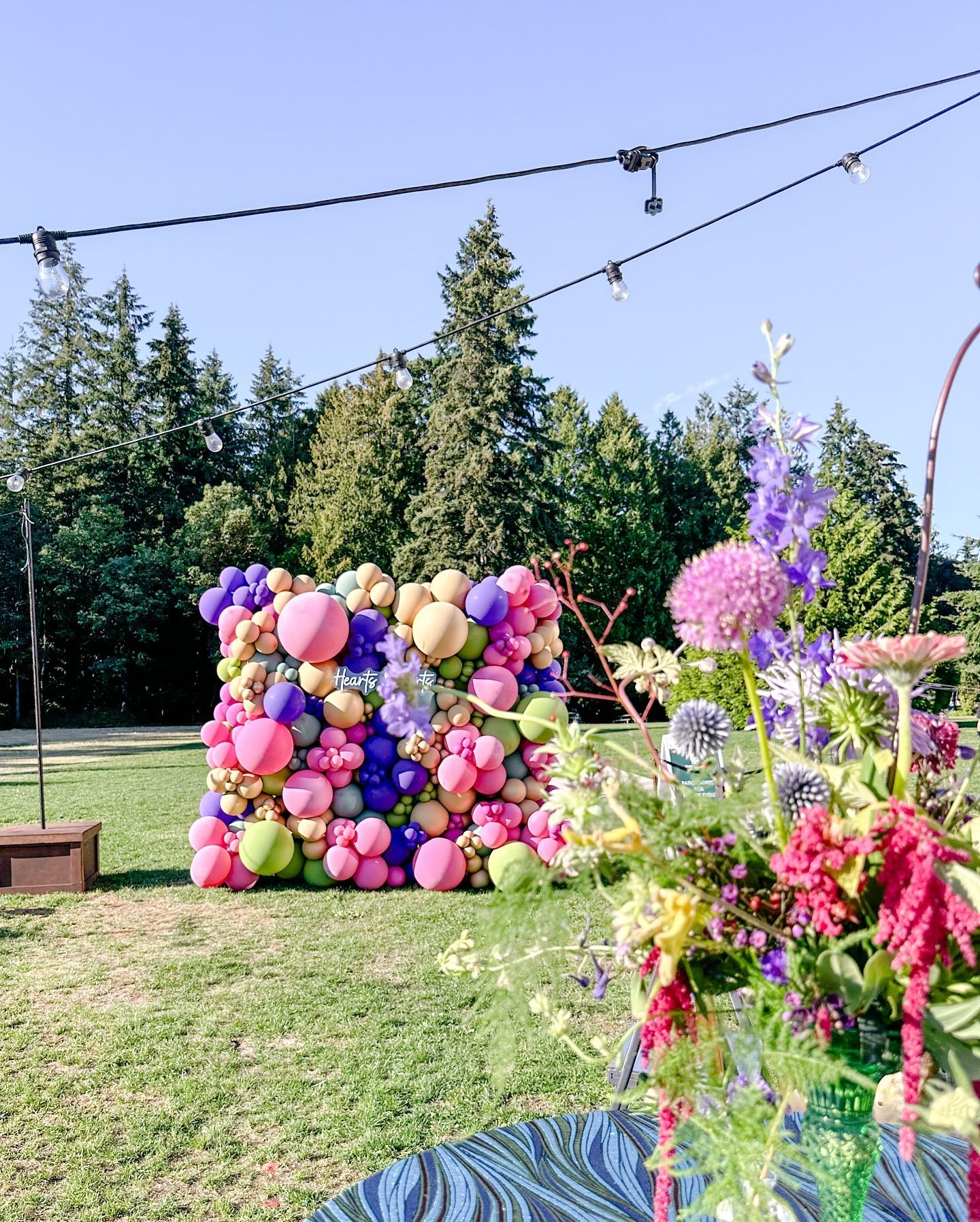 A wall of balloons and flowers in a field with trees in the background.