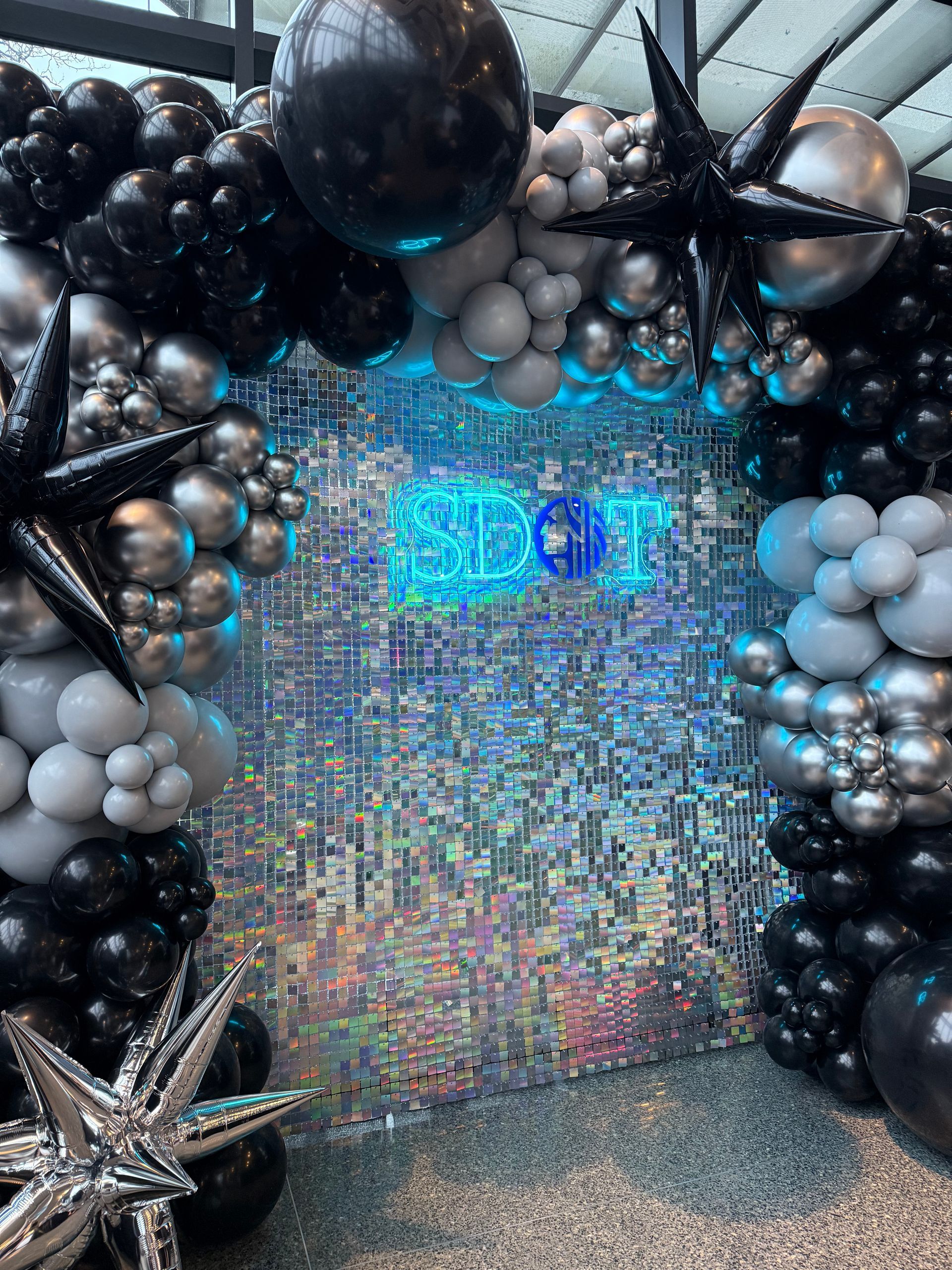Balloon arch with black, silver, and gray balloons framing a shimmering sequin wall with illuminated text.