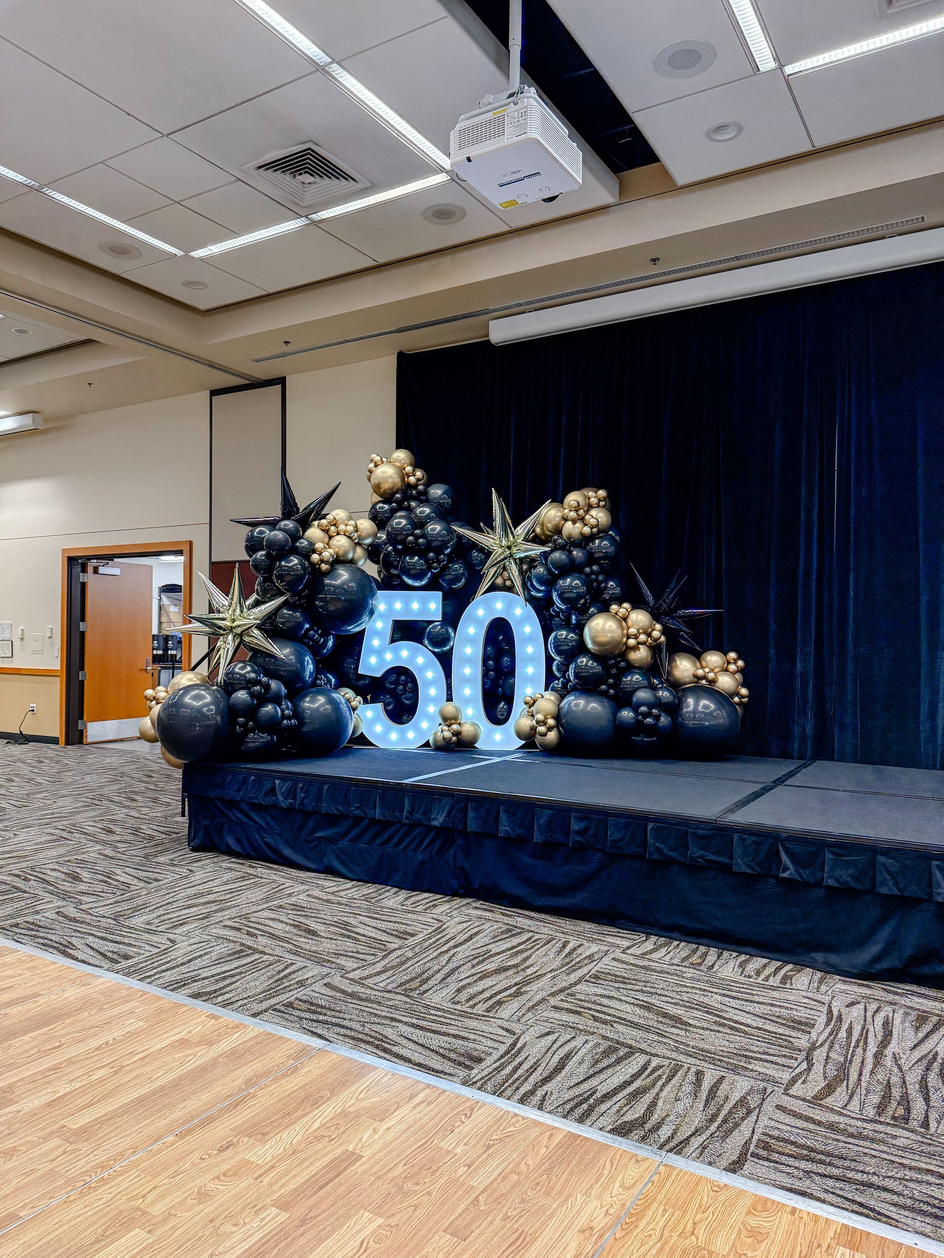 Stage decorated for a 50th celebration, with balloon arrangements in black and gold, and illuminated 
