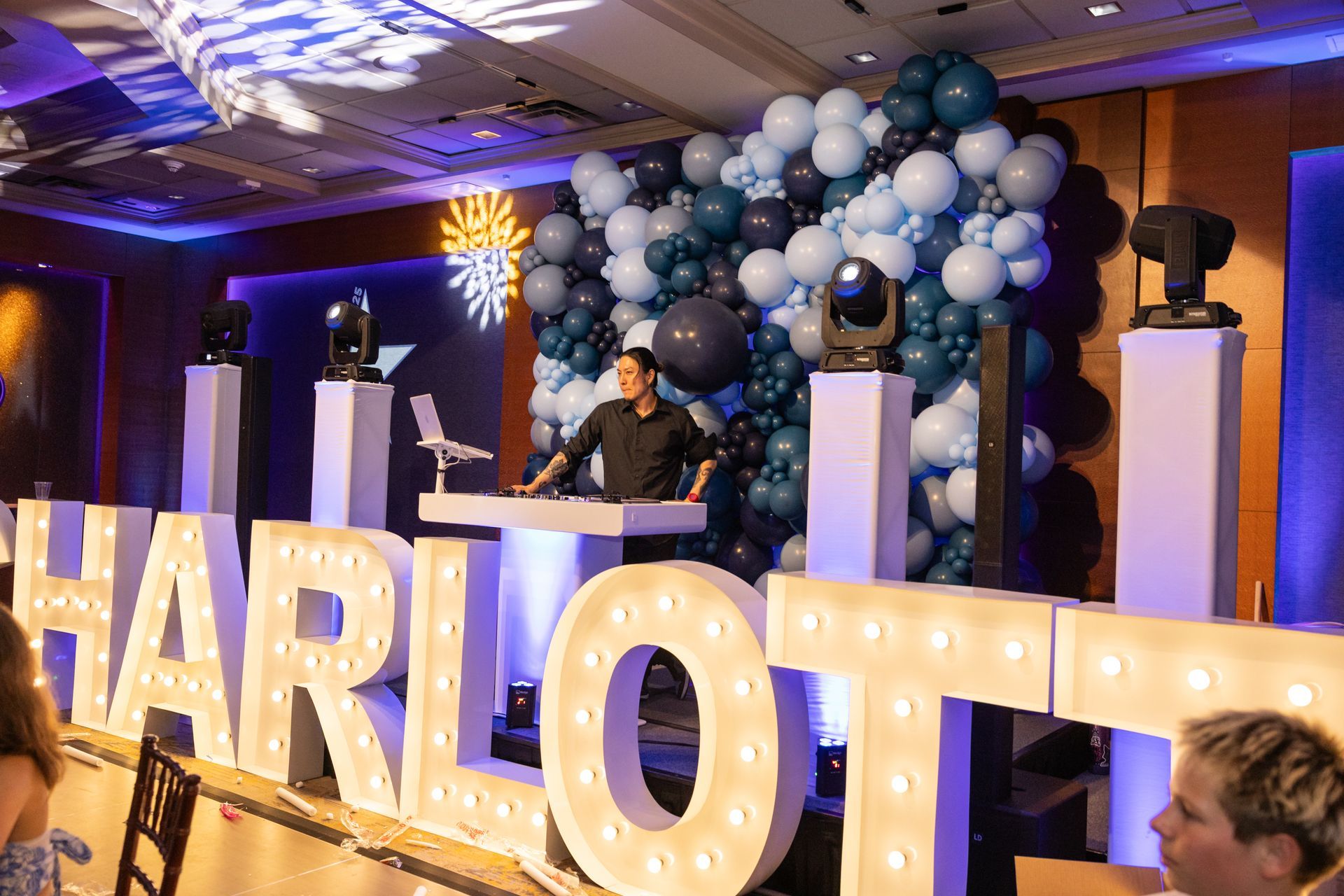 A person stands behind a DJ booth in front of a blue balloon wall and large, illuminated marquee letters spelling 
