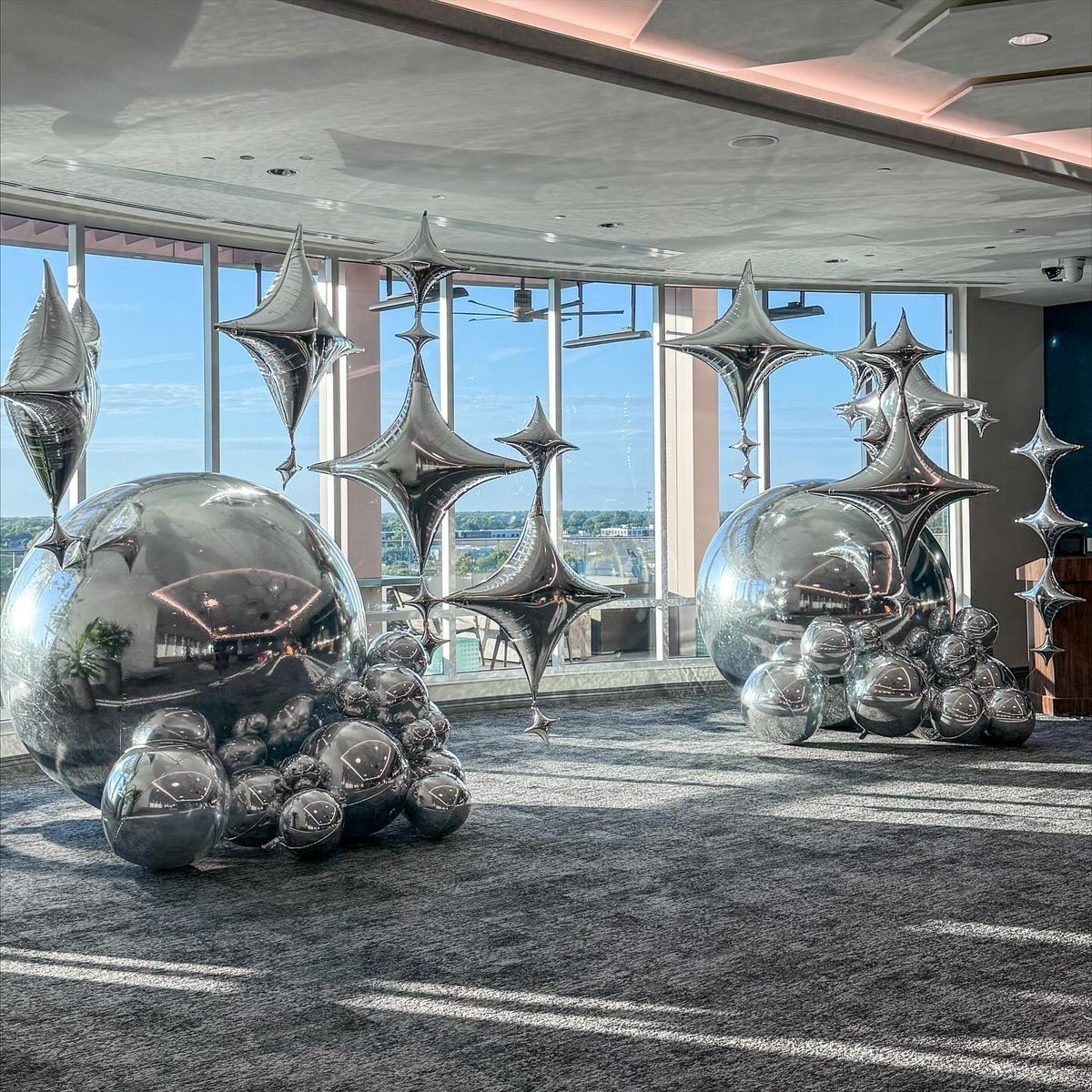 Silver geometric star-shaped sculptures hang above two clusters of metallic spheres on a gray carpeted floor by windows.