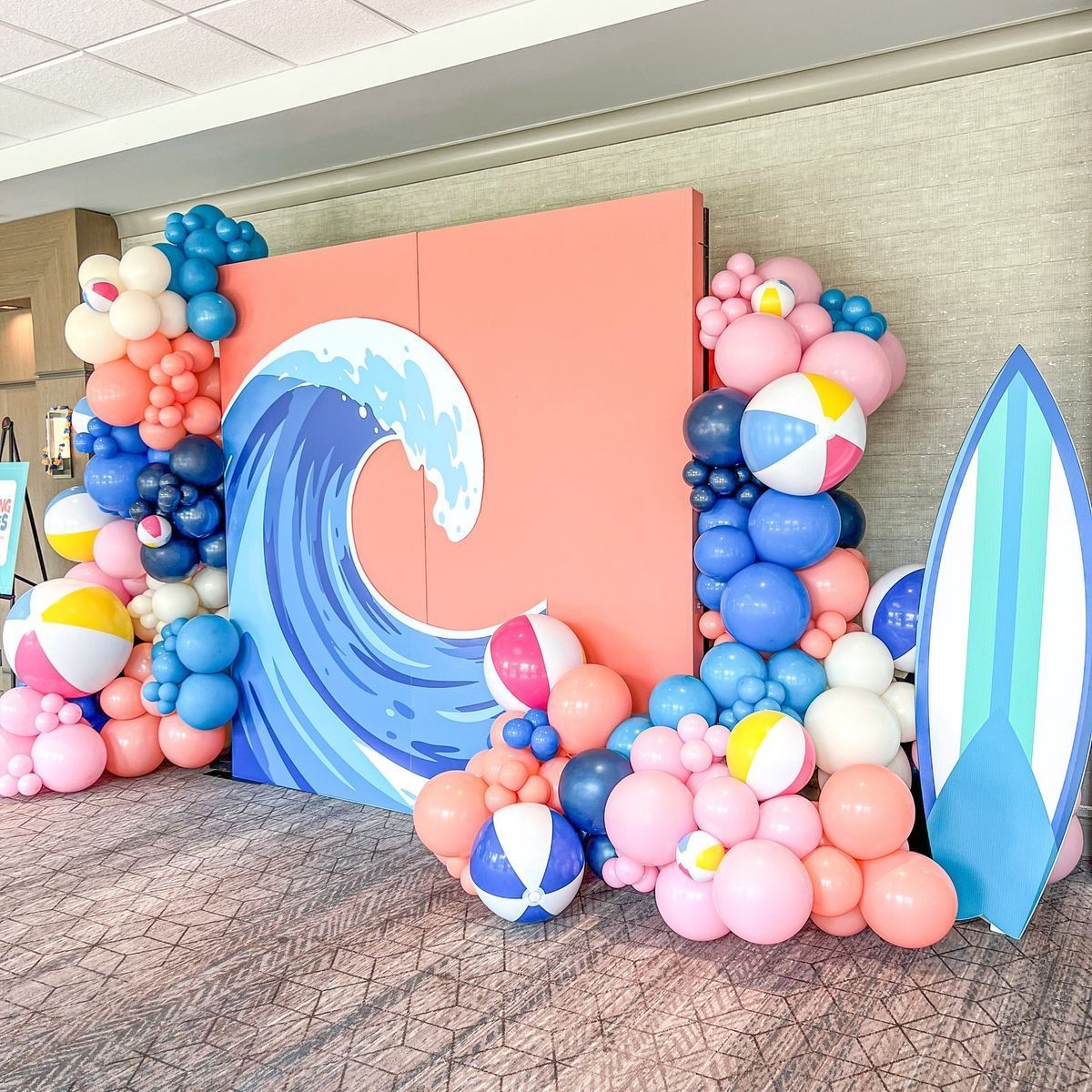 A beach-themed event backdrop featuring a coral-colored wall, a large wave graphic, colorful balloons, and a surfboard.