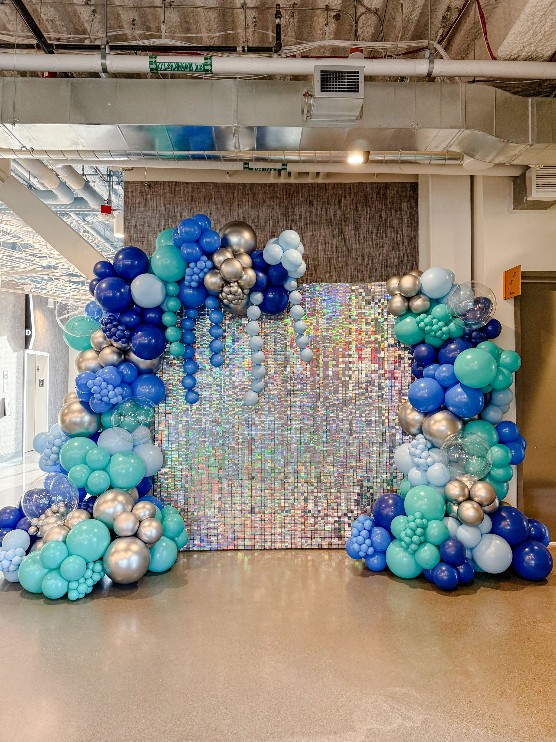Balloon arch with blue, teal, and silver balloons framing a sparkly backdrop, set indoors.