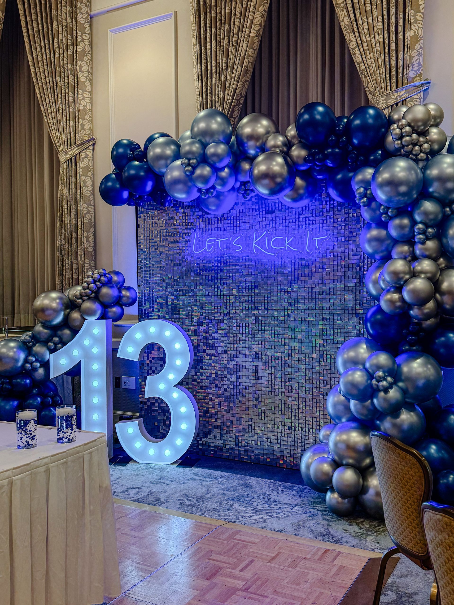 A decorated birthday display with a lighted number 13, blue neon sign, and a balloon arch in shades of blue and silver.