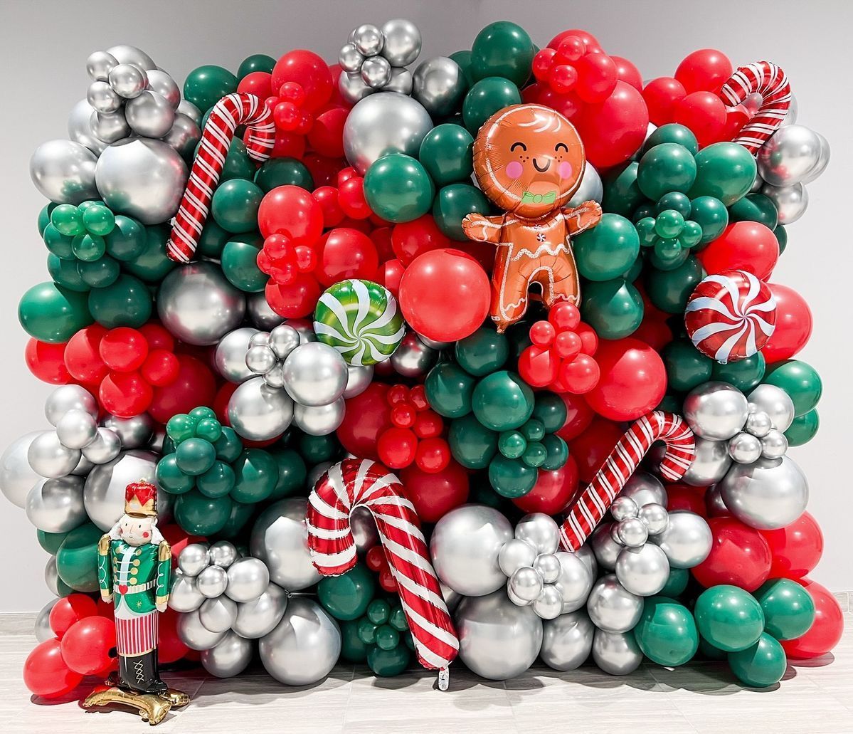 A festive Christmas balloon wall with red, green, and silver balloons, gingerbread man, candy cane, and nutcracker decor.